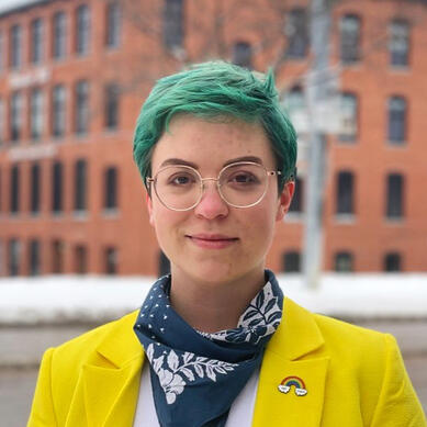 A person with green hair and glasses is looking into the camera and smiling. They are wearing a yellow jacket and a bandana